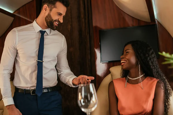 Избирательный фокус улыбающегося воздушного стюарда, смотрящего на счастливую, элегантную африканскую американку в частном самолете — стоковое фото