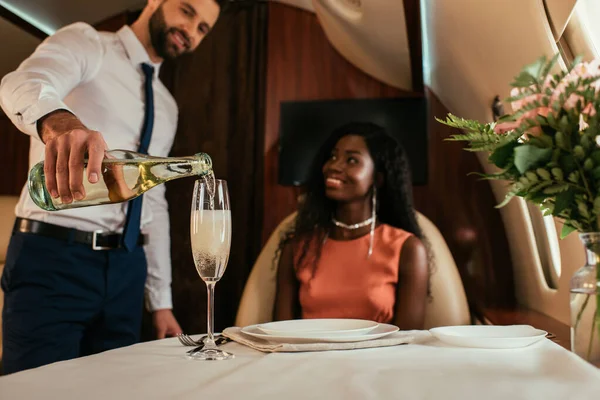 Foco seletivo do ar bonito mordomos derramando champanhe em vidro perto de mulher africana americana sorridente em avião privado — Fotografia de Stock