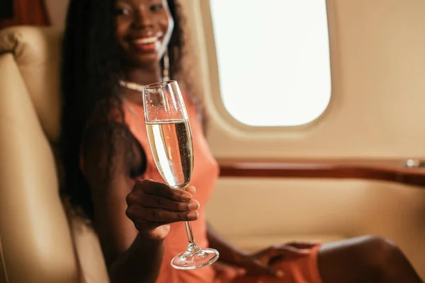 Selektiver Fokus einer glücklichen afrikanisch-amerikanischen Frau, die in die Kamera blickt, während sie ein Glas Champagner im Privatjet hält — Stockfoto