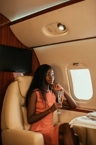 Elegant, dreamy african american woman holding glass of champagne and looking in porthole in private plane — Stock Photo