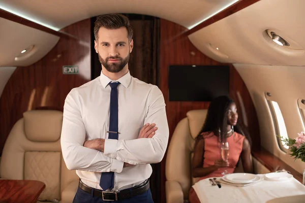 Handsome air steward with crossed arms looking at camera near elegant african american woman in private plane — Stock Photo