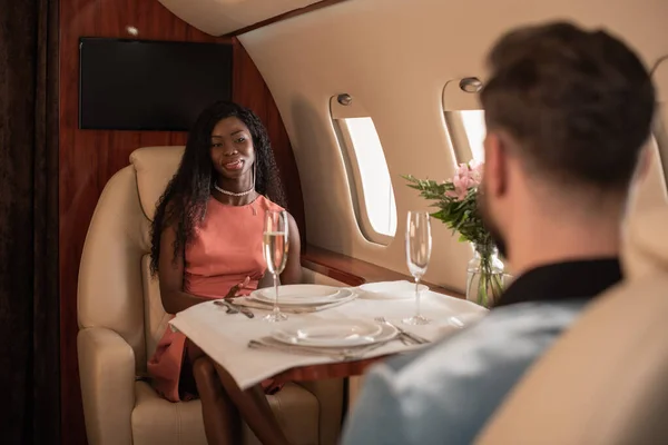 Rückansicht eines Mannes, der am gedeckten Tisch sitzt, gegenüber einer attraktiven, eleganten Frau im Privatflugzeug — Stockfoto