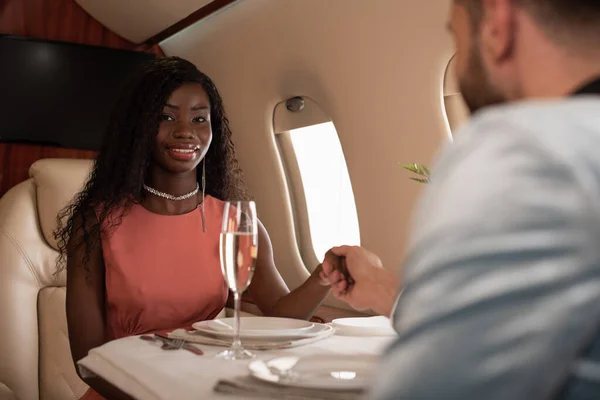 Foco seletivo de menina americana africana elegante olhando para a câmera enquanto segurando as mãos com o homem na mesa servida em avião privado — Fotografia de Stock