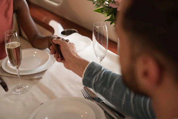 Partial view of interracial couple holding hands while sitting at served table in private plane — Stock Photo