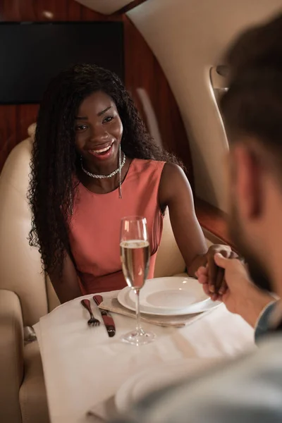 Foyer sélectif de heureuse femme afro-américaine tenant la main avec l'homme à la table servie dans un avion privé — Photo de stock