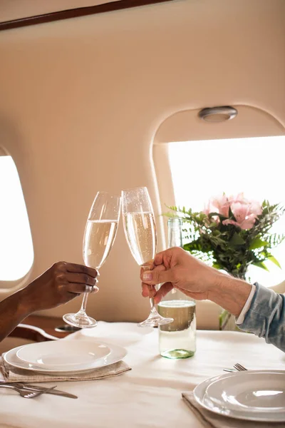 Teilansicht eines interrassischen Paares, das Champagnergläser an einem servierten Tisch im Privatflugzeug klimpert — Stockfoto