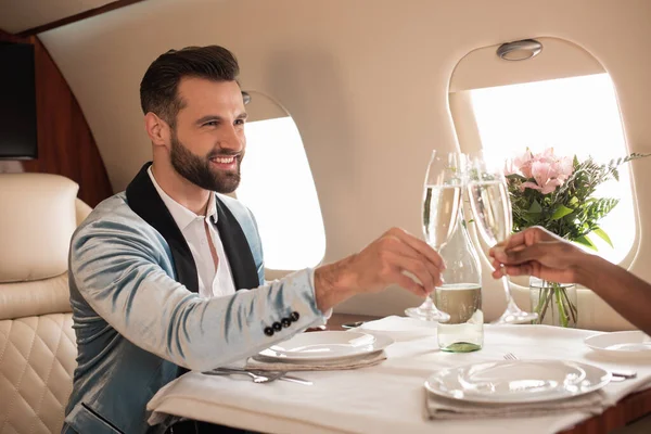 Vista parcial de la mujer afroamericana tintineo copas de champán con el hombre elegante en la mesa servida en avión privado - foto de stock