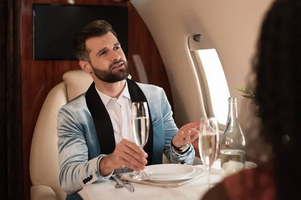 Abgeschnittene Ansicht einer afrikanisch-amerikanischen Frau, die an einem gedeckten Tisch im Privatflugzeug in der Nähe eines eleganten, sprechenden Mannes sitzt — Stockfoto