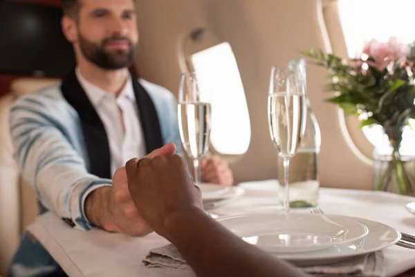 Selektiver Fokus eines glücklichen, eleganten Mannes, der Hand in Hand mit einer afrikanisch-amerikanischen Frau am gedeckten Tisch im Privatflugzeug hält — Stockfoto