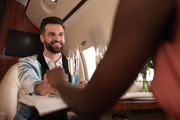 Abgeschnittene Ansicht einer afrikanisch-amerikanischen Frau, die Händchen hält mit einem lächelnden eleganten Mann am gedeckten Tisch im Privatflugzeug — Stockfoto