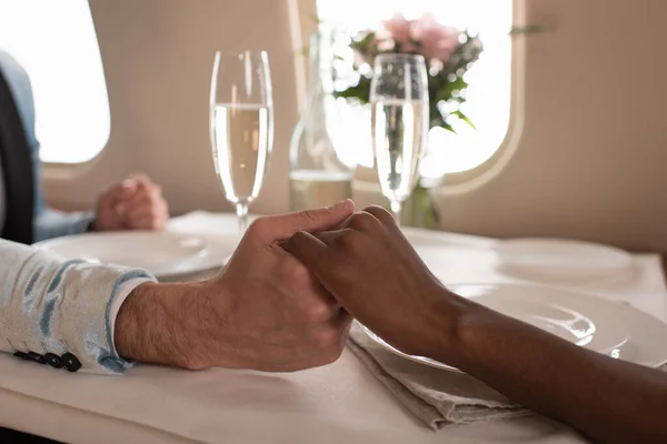 Vista cortada de casal interracial de mãos dadas perto de copos de champanhe na mesa servida em jato privado — Fotografia de Stock