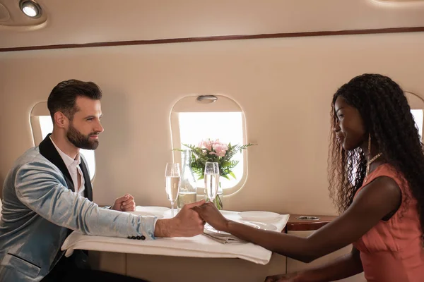 Happy, elegant interracial couple holding hands and looking at each other at served table in private jet — Stock Photo