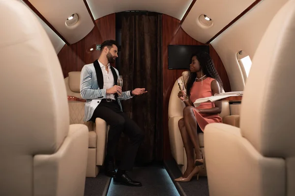 Избирательный фокус элегантной межрасовой пары, разговаривающей в частном самолете, держа бокалы для шампанского — стоковое фото