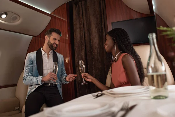 Selective focus of smiling interracial couple clinking glasses of champagne in private jet — Stock Photo