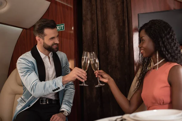 Happy, elegant interracial couple clinking glasses of champagne in private plane — Stock Photo