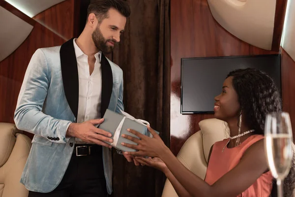 Handsome, elegant man presenting gift box to smiling african american woman in private plane — Stock Photo