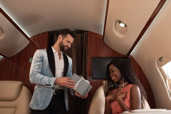 Bel homme élégant qui présente une boîte cadeau à une femme afro-américaine surprise dans un avion privé — Photo de stock