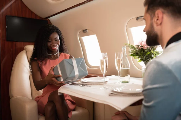 Selective focus of happy african american girl holding gift box near man and served table in private plane — Stock Photo