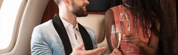 Vista recortada de pareja interracial tintineo copas de champán en avión, plano panorámico - foto de stock