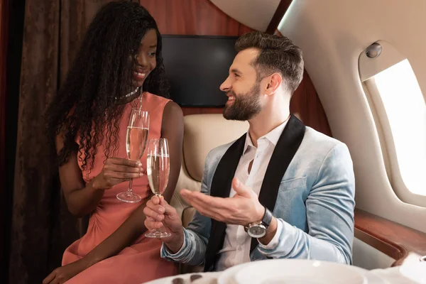 Улыбающаяся межрасовая пара путешествующая на частном самолете и разговаривающая, держа бокалы шампанского — стоковое фото
