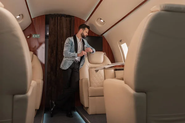 Selective focus of dreamy, elegant man holding glass of champagne and looking in porthole in private plane — Stock Photo