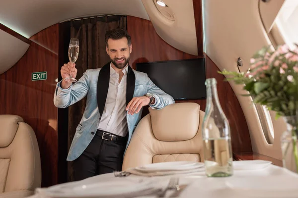Selektiver Fokus eines lächelnden, eleganten Mannes, der ein Glas Champagner in der Hand hält und im Privatflugzeug in die Kamera blickt — Stockfoto