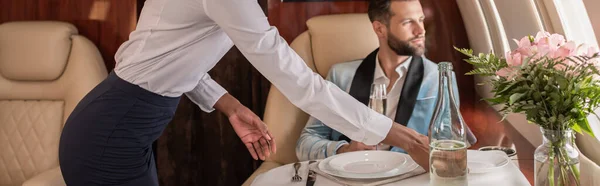 Vista ritagliata della hostess afro-americana apparecchiare la tavola in jet privato vicino all'uomo elegante, colpo panoramico — Foto stock