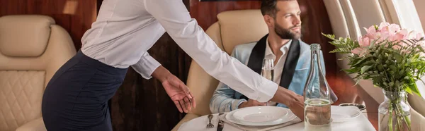 Vista cortada da mesa americana africana stewardess configuração em jato privado perto de homem elegante, tiro panorâmico — Fotografia de Stock