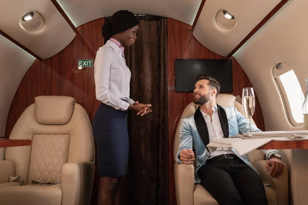 Atractivo, sonriente azafata afroamericana de pie cerca guapo, hombre elegante sentado a la mesa servida en jet privado - foto de stock