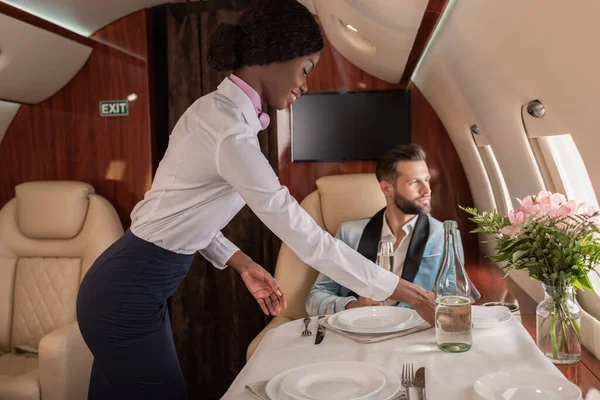 Sonriente, bastante afro americano azafata poner mesa en privado jet cerca elegante hombre - foto de stock
