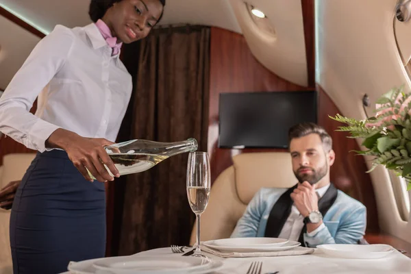 Selektiver Fokus attraktiver afrikanisch-amerikanischer Stewardess, die in der Nähe eines eleganten Mannes im Privatjet Champagner ins Glas gießt — Stockfoto