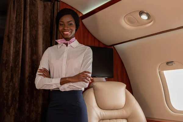 Attractive, cheerful african american stewardess standing with crossed arms in private jet and looking at camera — Stock Photo