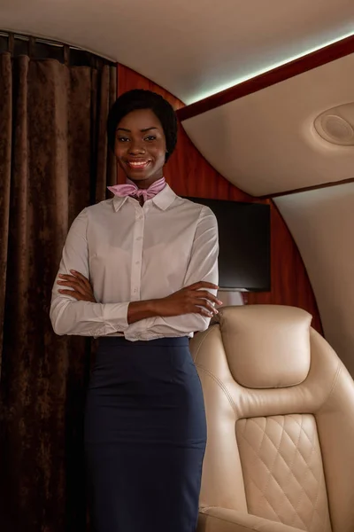 Beautiful, smiling african american stewardess standing with crossed arms in private jet and looking at camera — Stock Photo