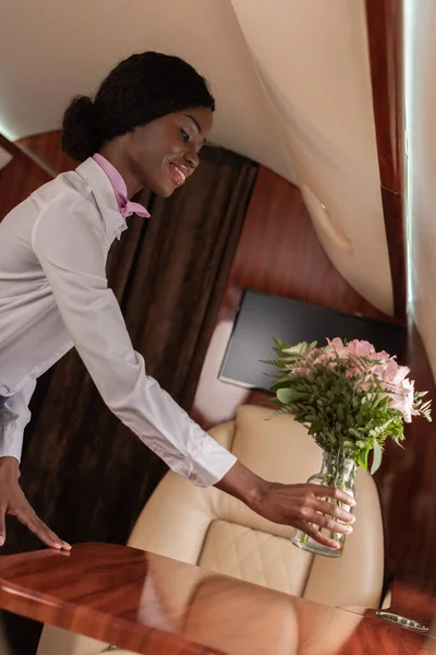 Smiling african american stewardess putting fresh flowers on table in private jet — Stock Photo