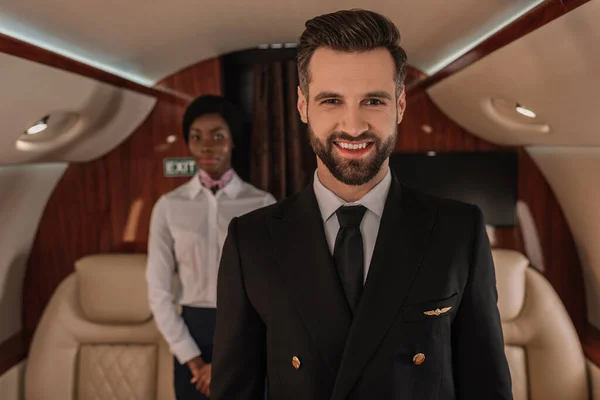 Enfoque selectivo de piloto guapo sonriendo a la cámara cerca de azafata afroamericana - foto de stock