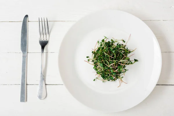 Вид сверху на свежий микрозеленый цвет на тарелке возле столовых приборов на белой деревянной поверхности — стоковое фото