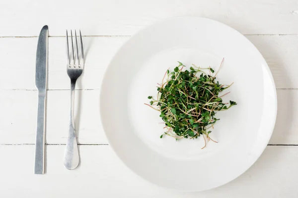 Vista superior de microgreen fresco en la placa cerca de cubiertos en la superficie de madera blanca - foto de stock