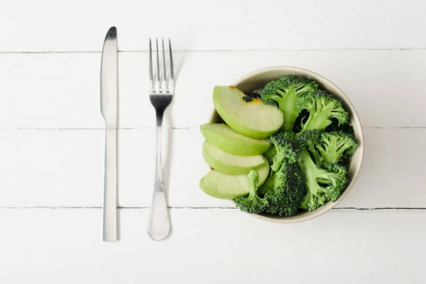 Top view of fresh green apple and broccoli in bowl near cutlery on white wooden surface — Stock Photo