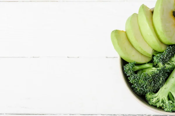 Vista superior de la manzana verde fresca y el brócoli en un tazón sobre una superficie de madera blanca - foto de stock