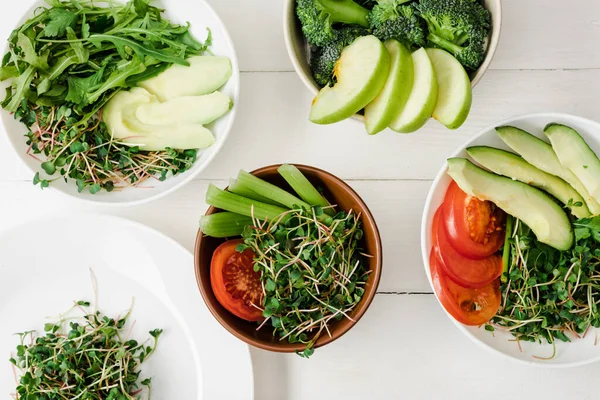 Top view of fresh vegetables and fruits with microgreen in bowls on white wooden surface — Stock Photo