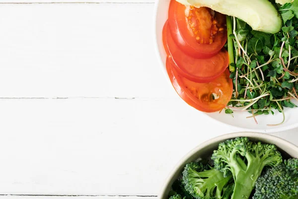 Vista superior de microgreen fresco, tomate, abacate e brócolis em tigelas na superfície de madeira branca — Fotografia de Stock