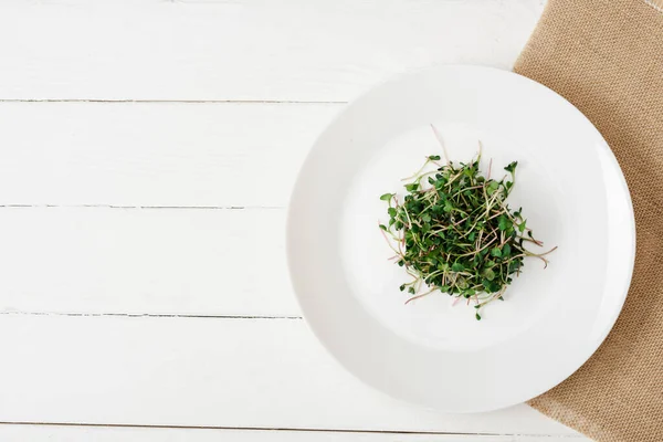 Vista superior de microgreen fresco na placa no guardanapo bege na superfície de madeira branca — Fotografia de Stock