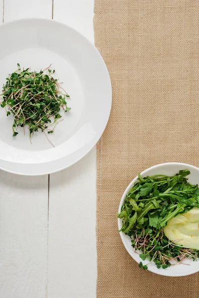 Vista superior do microgreen fresco na placa perto da tigela da salada verde no guardanapo bege na superfície de madeira branca — Fotografia de Stock