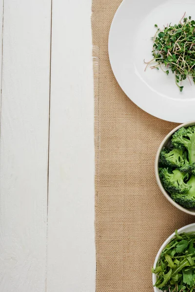 Vista superior de microgreen fresco na placa perto de tigelas de legumes verdes no guardanapo bege na superfície de madeira branca — Fotografia de Stock