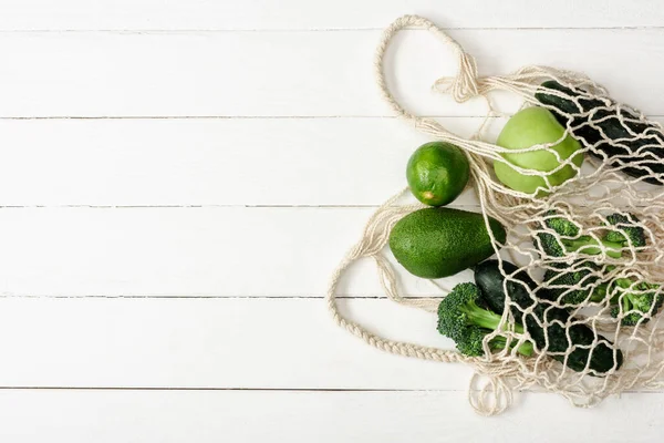 Vista superior de frutas y verduras verdes frescas en bolsa de hilo sobre superficie de madera blanca - foto de stock