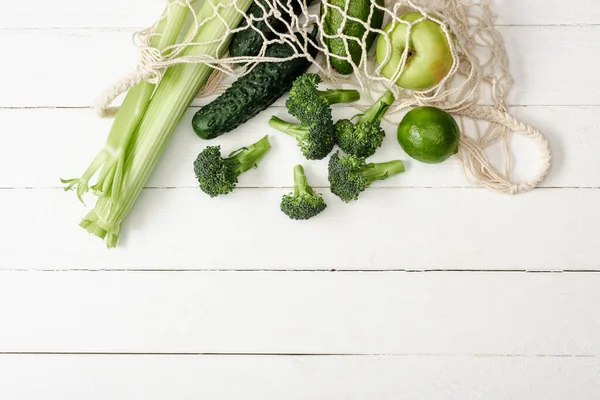 Top view of fresh green fruits and vegetables in string bag on white wooden surface — Stock Photo