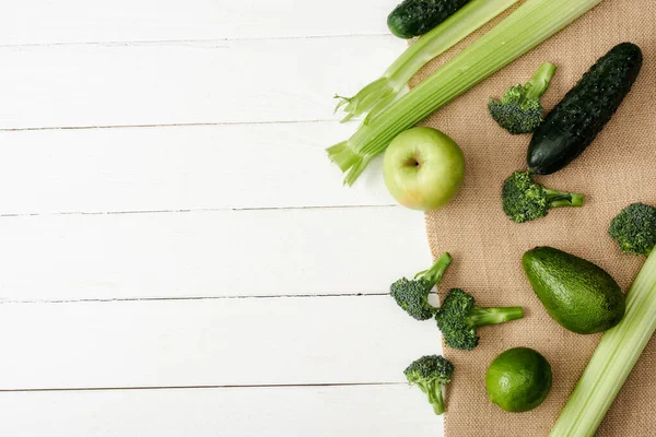 Vista superior de frutas y verduras verdes frescas sobre tela de saco sobre superficie de madera blanca — Stock Photo