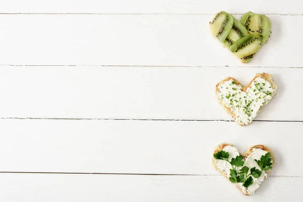 Vue de dessus de la canape en forme de coeur avec fromage crémeux, microvert, persil et kiwi sur la surface en bois blanc — Photo de stock