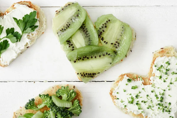 Vista superior de canapé em forma de coração com queijo cremoso, brócolis, microverde, salsa e kiwi na superfície de madeira branca — Fotografia de Stock