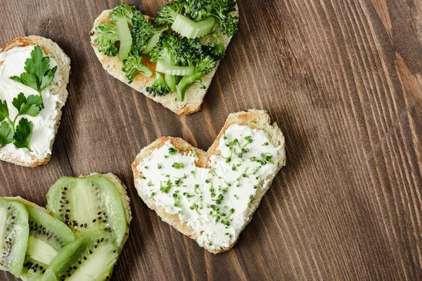 Vista superior de canapé em forma de coração com queijo cremoso, brócolis, microverde, salsa e kiwi na superfície de madeira — Fotografia de Stock